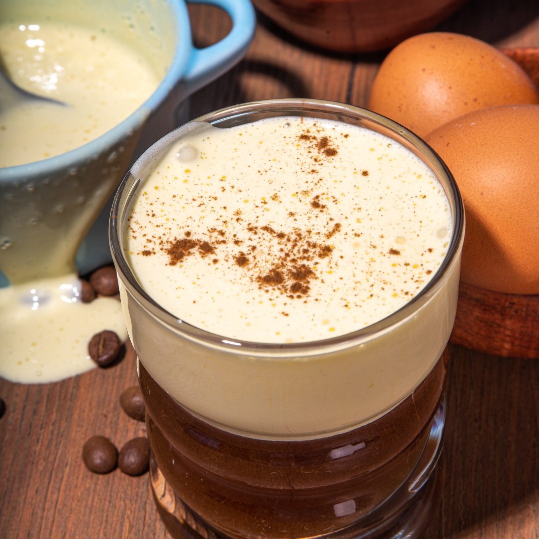A steaming cup of Vietnamese Egg Coffee with a thick, creamy golden egg foam on top, served in a glass cup on a wooden table. The rich, frothy texture contrasts beautifully with the dark coffee beneath, creating a layered, indulgent drink.